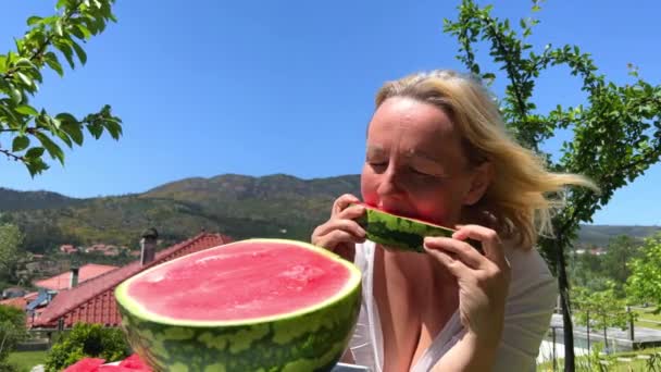 Uma mulher adulta gananciosamente come uma melancia, morde muitas vezes, flui pelos braços e ela está feliz com o pano de fundo das montanhas e uma grande melancia — Vídeo de Stock