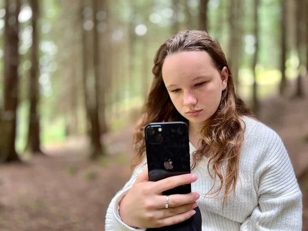 Dívka v bílé košili teenager leží na kameni v rukou drží černý iPhone 13 Dívá se na obrazovku klidně zvedá ruce — Stock fotografie