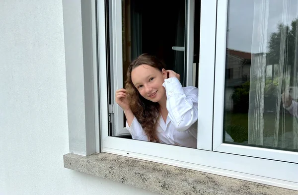 Una bella ragazza adolescente guarda fuori da una finestra bianca è in una camicia bianca sorridente e molto felice e felice ride — Foto Stock