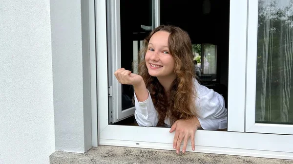 漂亮的少女朝窗外看去，挥挥挥手吸引别人的注意。她穿着一件白色衬衫，房子的窗户是白色的 — 图库照片