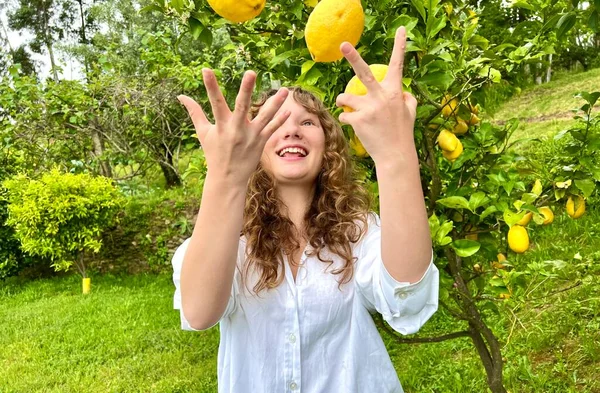 Una chica de pelo rubio hace malabares con limones contra el telón de fondo de un limonero en un país tropical, está vestida con una camisa blanca se ríe y se divierte —  Fotos de Stock