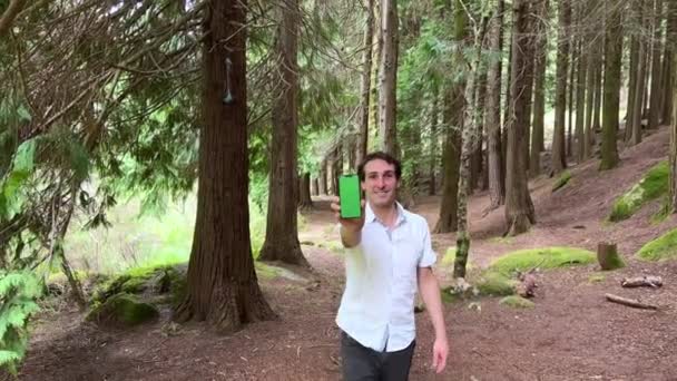 Pohledný španělský muž ukazuje telefon, na kterém stojí zelený displej chromakey v lese v bílé košili — Stock video