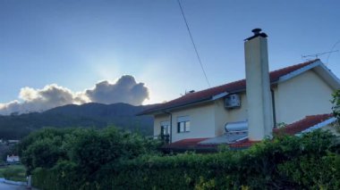 Dağlarda kırmızı çatılı beyaz bir ev dağın tepesindeki büyük bir buluta karşı güneş bulutları aydınlatıyor.