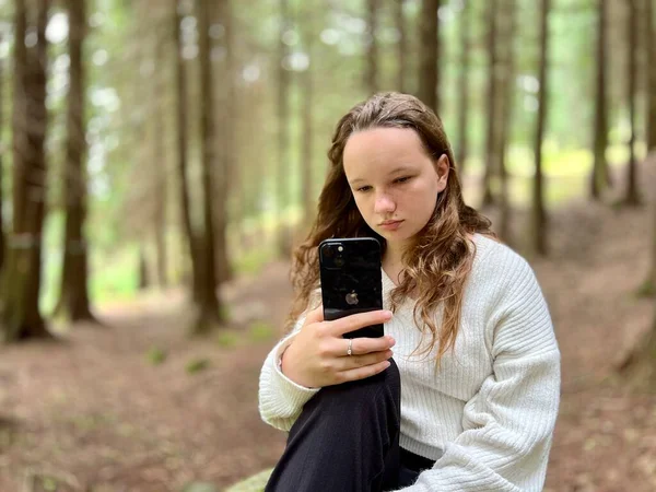 En flicka i vit skjorta en tonåring ligger på en sten i hennes händer hon håller en svart iPhone 13 Hon tittar på skärmen lugnt upp händerna — Stockfoto