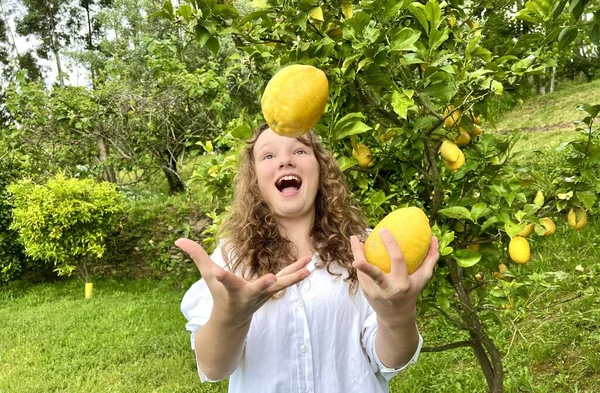 Una chica de pelo rubio hace malabares con limones contra el telón de fondo de un limonero en un país tropical, está vestida con una camisa blanca se ríe y se divierte —  Fotos de Stock