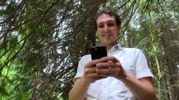 Un hombre de apariencia española está de pie en el bosque con un teléfono iPhone 13 negro, que está mirando el teléfono en una camisa blanca sin mirar hacia arriba desde la pantalla — Vídeos de Stock