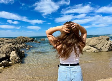 Kumsalda kot pantolonlu ve tişörtlü genç bir kız denizde oturur saçlarını düzleştirir ve gözlerini kapayıp başını göğe kaldırır.