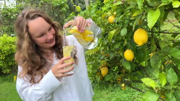 レモンの木を背景に10代の少女がレモンを飲みながらガラスのレモネードを片手に酒の瓶を持ち — ストック動画