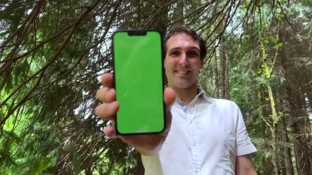 手机上拿着绿色屏幕彩色钥匙 拿着一个男人的手机 他站在森林里 穿着一件白色衬衫 把手机给摄像机看 高质量的4K镜头 — 图库视频影像