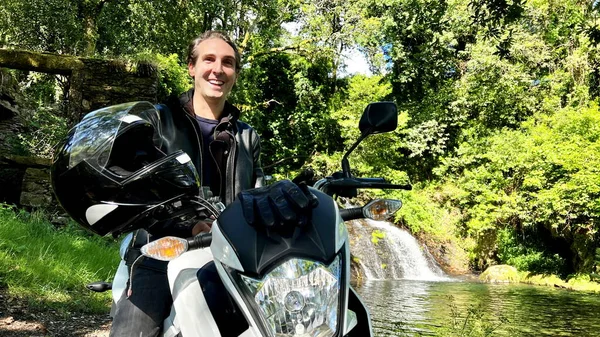 a handsome French man on a motorcycle against the backdrop of a landscape with a waterfall in Portugal he is in a black jacket he has an expressive face and a smile. High quality photo
