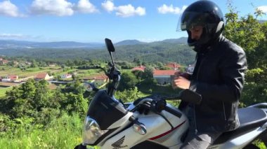 Siyah ceketli ve kafasında motosiklet kaskı olan bir adam dağların arka planında ve Portekiz 'de beyaz bulutlu mavi bir gökyüzü eldivenlerini giyer. Yüksek kalite 4k