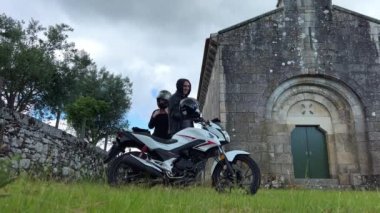 Motosikletin yanına kask takmış bir adam ve bir kadın. Terk edilmiş eski bir kilisenin arka planında duruyorlar ve gri gökyüzü, siyah giyinmişler ve motosiklet beyaz.