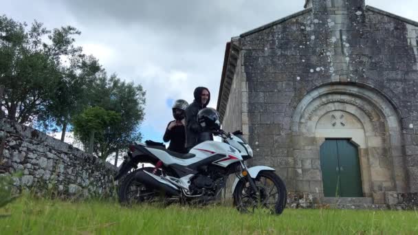 一个男人和一个女人在摩托车旁边戴上了钢盔 他们站在一座废弃的旧教堂和灰色的天空的背景下 身穿黑色衣服 摩托车是白色的 — 图库视频影像