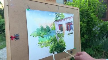 Yetişkin erkek sanatçı 45-55 yaşlarında bir resim çiziyor. Önündeki eve bakıyor. Üzerinde pencereleri, yeşil ahşabı ve bir sürü fırçayı görebileceğiniz bir kağıt parçası..