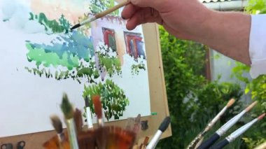 Yetişkin erkek sanatçı 45-55 yaşlarında bir resim çiziyor. Önündeki eve bakıyor. Üzerinde pencereleri, yeşil ahşabı ve bir sürü fırçayı görebileceğiniz bir kağıt parçası..