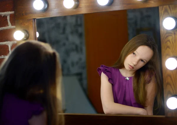 child girl looks in mirror with lights in purple dress and place for text wooden mirror in bedroom. High quality photo