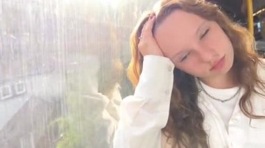 European girl with blond long hair in a white shirt rides in a skytrain. she falls asleep holding her head with her hand she is sad a little sad maybe she is tired of the long journey every day