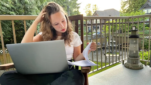 Online learning fair-haired european girl sits on her knees with a laptop on the street on the balcony or watches a lesson printouts of sheets on which she will do task