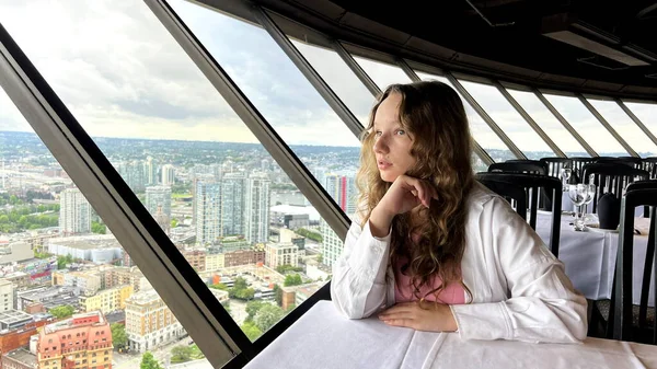 a young teenage girl sits at the window of a revolving restaurant and is sad she is sad her hand near her face and she looks out the window at the sights Top Of Vancouver Revolving Restaurant. 4k