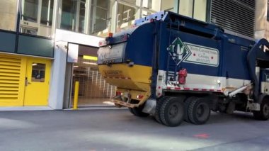 Çöp kamyonu Vankuwea 'nın merkezindeki sokaktan ayrılır. Geri geri gider ve caddede döner. Sarı ve beyazla birlikte mavi renktedir. Yüksek kaliteli 4K görüntüler Vancouver Canada 07.2022