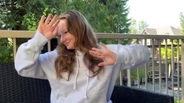 a teenage girl in a gray sweatshirt is dancing on the terrace, she has an oversized hoodie she has white wireless headphones in her ears. she smiles her light-colored hair is loose outdoors summer.