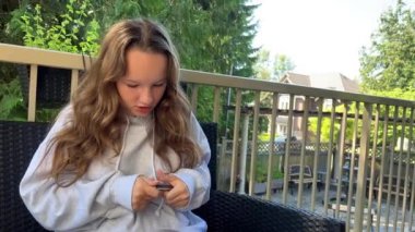 A girl in a gray hoodie is trying to take off her phone case. She is looking at him intently. She has blond flowing hair behind. Summer and green leaves. Maybe 8. High quality 4k footage