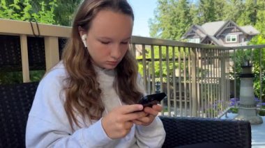 A teenage girl in a gray hoodie sits on the veranda on the terrace and looks at the phone she is busy She has a black iPhone 13 in her hands trees behind her and it is summer. High quality 4k footage