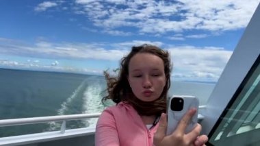 a teenage girl with blond hair in a pink sweater takes a selfie against the backdrop of the ocean she stands on a liner that sails in the Pacific Ocean and looks at herself in camera of a mobile phone