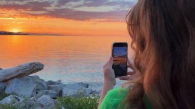 a teenage girl in a Green T-shirt stands with her back to the camera and shoots a beautiful sunset on Pacific Ocean she holds an iPhone 13 phone in her hands blond red hair stones setting sun sea sky