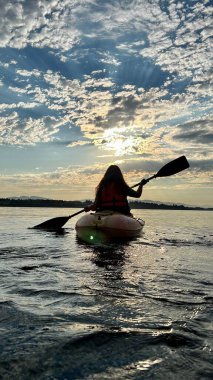 a teenage girl is kayaking at sunset in Pacific Ocean, only the silhouette of Kayak and Paddles is visible she swims along a sunny path to the sea. a beautiful sky with small clouds 08.2022 Canada 