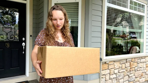 The daughter went out onto the porch, picked up the delivery of a cardboard box and looks around, she wonders who brought the delivery, no one is visible on the street.