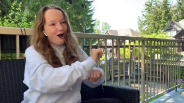 a teenage girl in a gray sweatshirt is dancing on the terrace, she has an oversized hoodie she has white wireless headphones in her ears. she smiles her light-colored hair is loose outdoors summer.