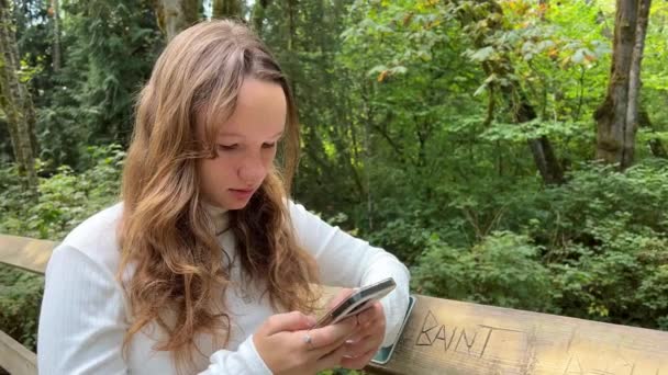 白いタートルネックの女の子は自然の中で電話で忙しいです自然夏の公園の少女ティーネージャーサリーのTynehead地域公園ソーシャルネットワークのティーネージャーの生活の中で — ストック動画