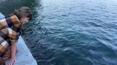 Stanley Park Pasifik Okyanusu kıyısında oturan yetişkin bir kadın ve suya bakıyor, güneş gözlüğü ve turuncu elbiseler giyiyor kahverengi ekose ceket ve deri etek giyiyor sonbahar ılık havası