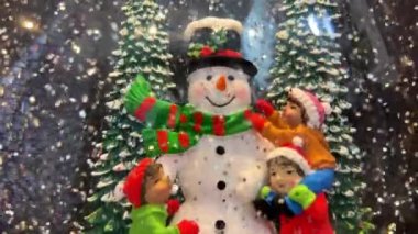 Kar küresi. Kardan bir adam, yanında iki Noel ağacı arasında duruyor. Üç çocuk, ona yeni inşa etmiş.
