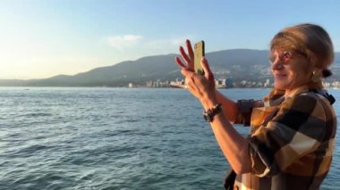 Ekose kahverengi ceketli yetişkin bir kadın elinde telefon ve saatle elinde bir kadın iPhone Stanley Park Vancouver Canada 'da arka planda dağlarla video çekiyor.
