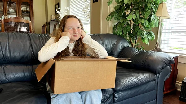 A teenage girl sits sadly near an open parcel and looks straight at the box, leaning on her elbow, on which you can write something if there is a place for text. She is in White Golf in the room
