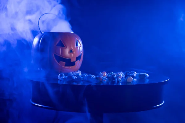 A plastic pumpkin jack o lanter bucket for trick or treating with saltwater taffy on a table against a back background with fog