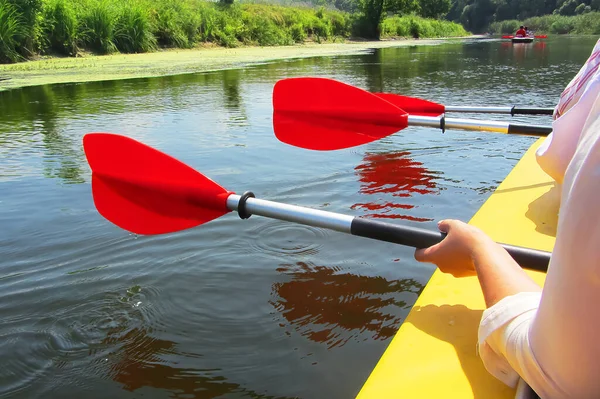 Resimde: Kano üzerinde yüzen insanların ellerinde peş peşe üç kürek çekiliyor. Kürekler sakin suya yansıyor. Sarı kanodaki yüzen insanların elinde kırmızı raketler var.