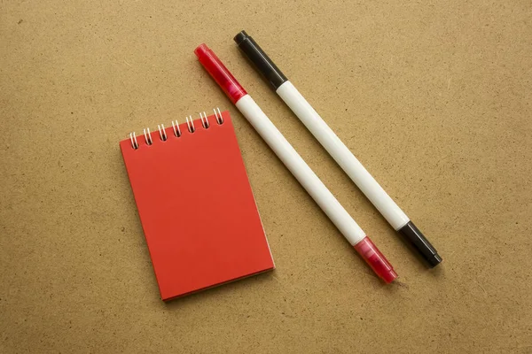 Red notepad for notes with red sheets. Blank space for notes. Mocap. Two felt-tip pens in red and black. On a warm ochre gray background. Holiday calendar notes on notepad. Background for inscriptions