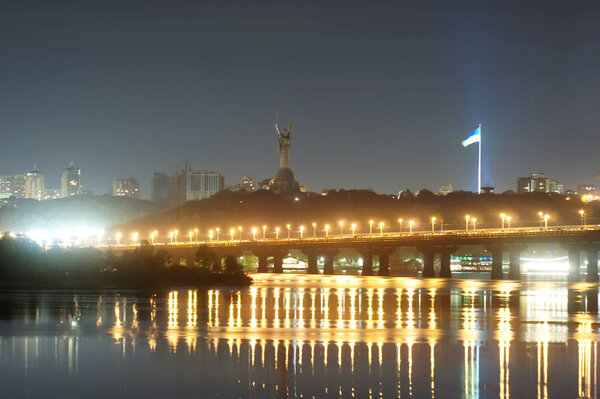 Beautiful view of Kiev, the capital of Ukraine. Night view across the Dnieper River. The lights of the city at night are reflected in the river. View of the flag.