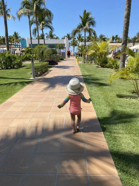 Toddler walking down pathway on vacation at resort in sun hat 