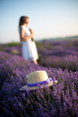 A straw hat with violet tape lays on a top of lavender flowers, the unfocused person behind the main object