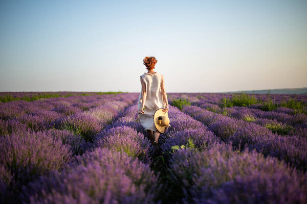 Young woman holding a straw hat in her hands running on the lavender field. Girl dressed in a linen dress and a straw hat running between lavender flowers. Back view. Horizontal photo