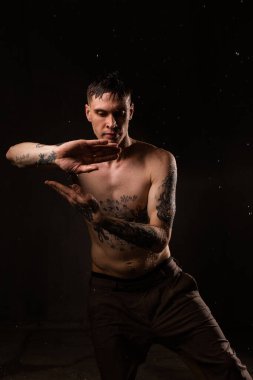 Vücudunda su damlaları olan dövmeli bir adam yağmurda yakışıklı, sağlıklı dövmeli genç bir adam. Seksi fitness adamı. Dövmeli çıplak gövde. Moda erkeği stüdyoda karanlık arka planda poz veriyor..