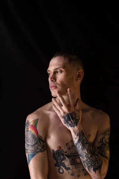 Tattooed man with water drops on his body, handsome healthy tattooed young man in the rain. Sexy fitness man. Naked torso with tattoos. Fashion male posing in studio on dark background.
