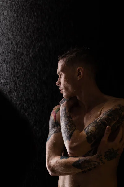 Tattooed man with water drops on his body, handsome healthy tattooed young man in the rain. Sexy fitness man. Naked torso with tattoos. Fashion male posing in studio on dark background.