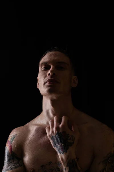 Tattooed man with water drops on his body, handsome healthy tattooed young man in the rain. Sexy fitness man. Naked torso with tattoos. Fashion male posing in studio on dark background.