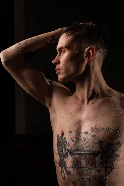 Tattooed man with water drops on his body, handsome healthy tattooed young man in the rain. Sexy fitness man. Naked torso with tattoos. Fashion male posing in studio on dark background.