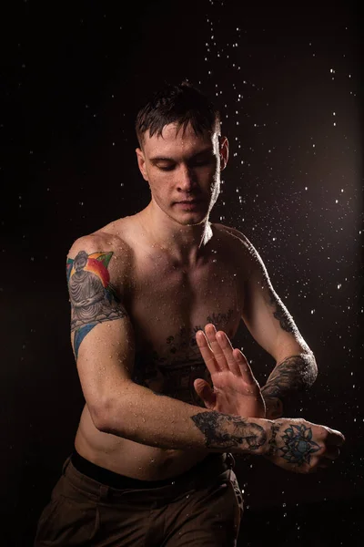 Tattooed man with water drops on his body, handsome healthy tattooed young man in the rain. Sexy fitness man. Naked torso with tattoos. Fashion male posing in studio on dark background.